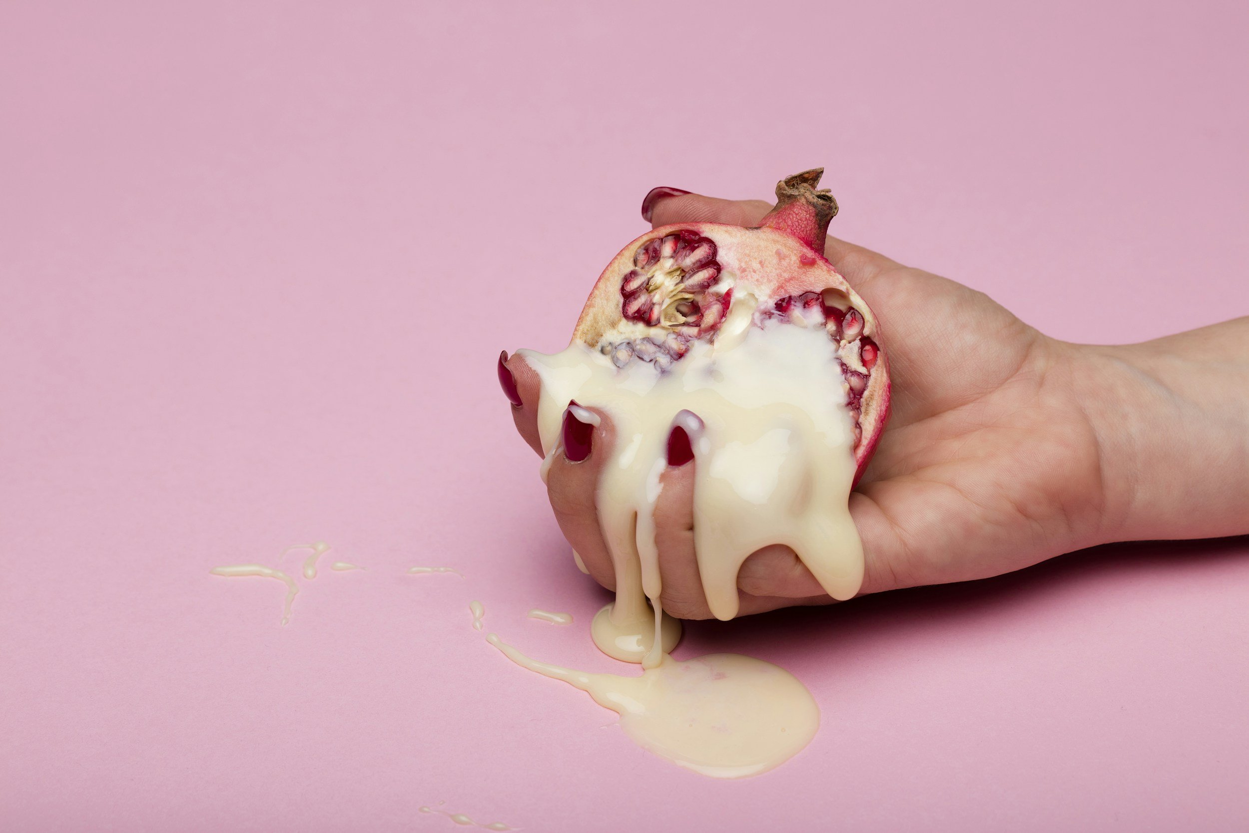 A hand holding a halved pomegranate with white cream or sauce dripping from it onto a pink background.