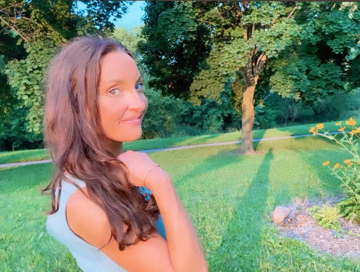 A woman with long brown hair and a sleeveless top standing in a park with green grass, trees, and sunlight casting shadows.
