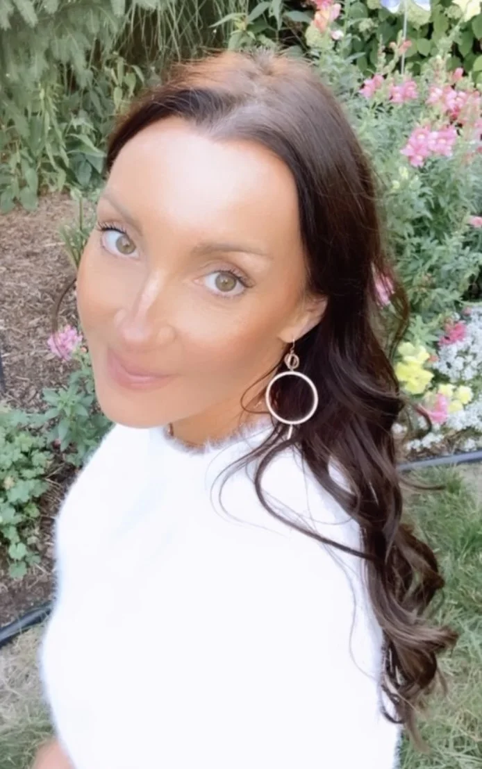 Woman with long wavy brown hair wearing circular earrings, smiling, outdoors in a garden with pink flowers and green foliage.