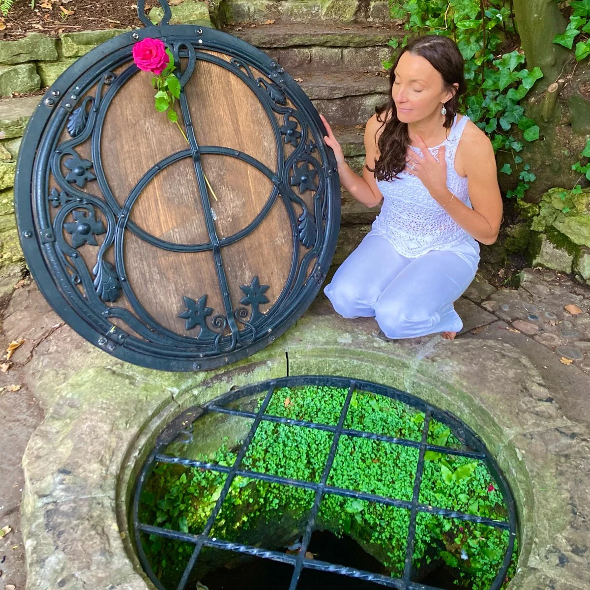 A woman dressed in white kneels beside an open decorative metal cover over a well filled with green plants, with a pink rose resting on the cover.