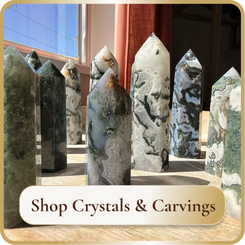 A collection of carved and polished crystals on a wooden surface inside a shop, with sunlight coming through a window. The sign reads "Shop Crystals & Carvings."