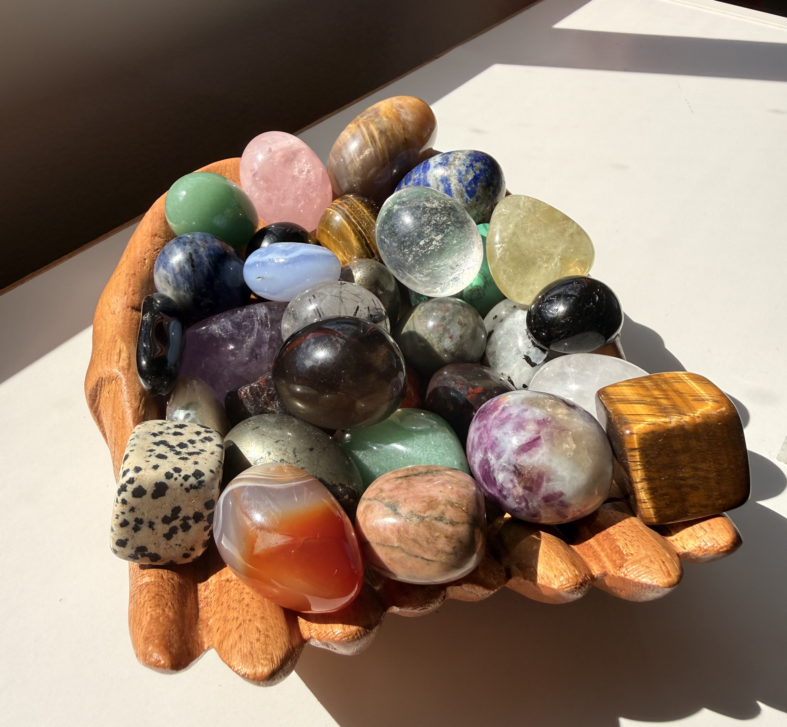 A wooden bowl filled with various polished gemstones and minerals, placed on a white surface. Sunlight casts shadows and highlights on the stones.