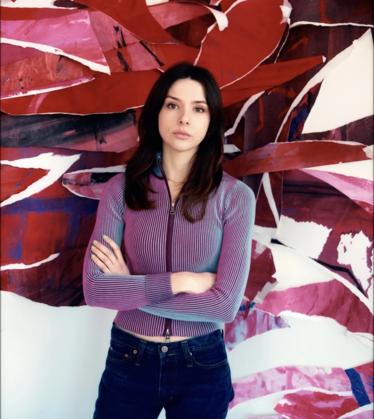 A woman with long hair standing in an art studio, smiling, with multiple abstract paintings on the floor and wall behind her, under a sloped wooden ceiling with skylights.