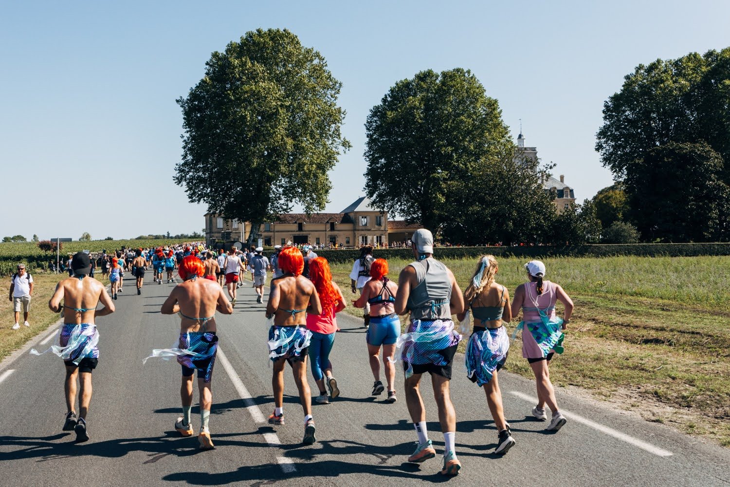 Group of people participating in an outdoor parade or event, some wearing colorful costumes and wigs, walking on a road with trees in the background.
