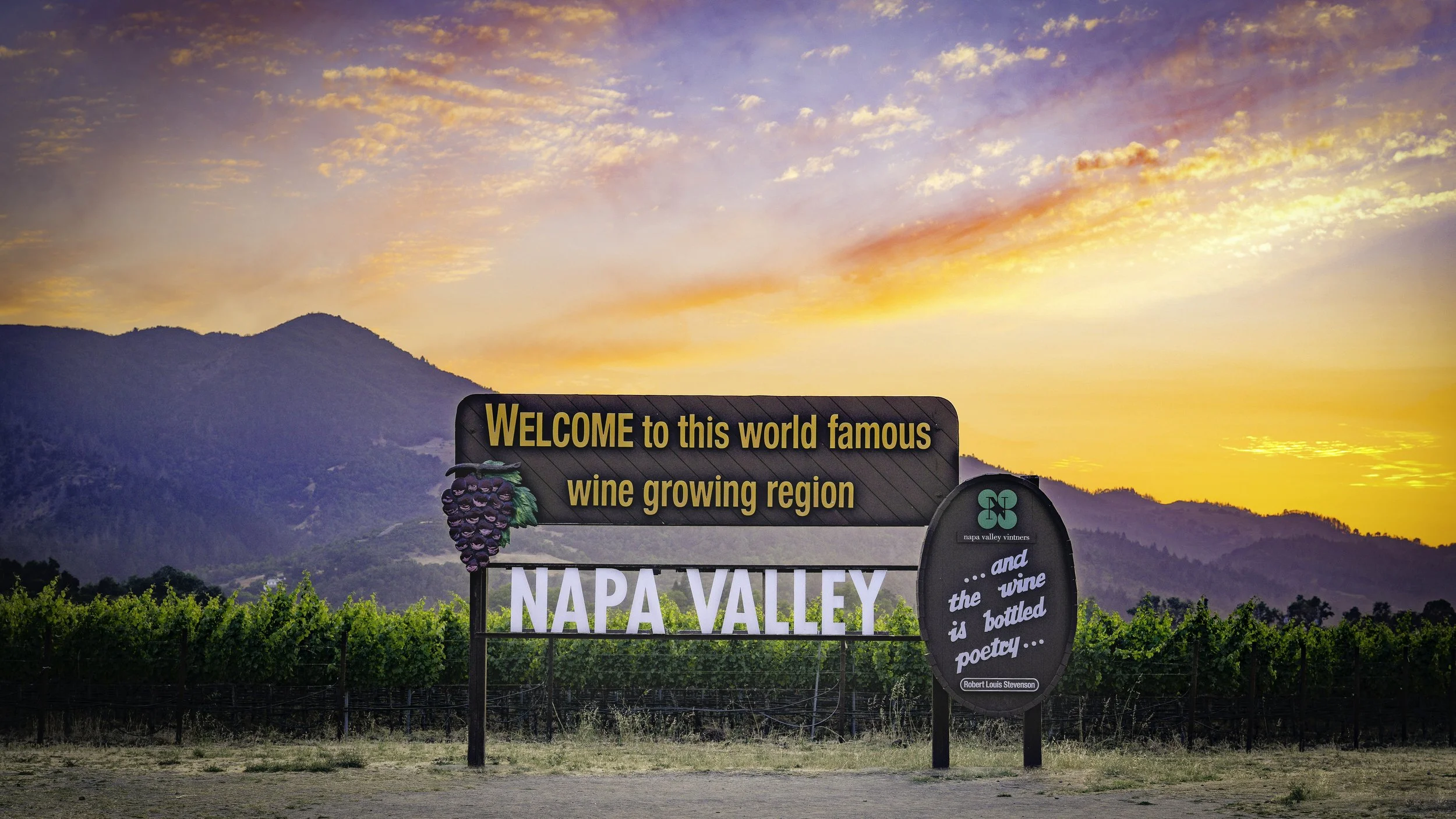 Sign welcoming visitors to Napa Valley, a famous wine-growing region, with mountains and a sunset sky in the background. The sign features grapes and quotes about wine.