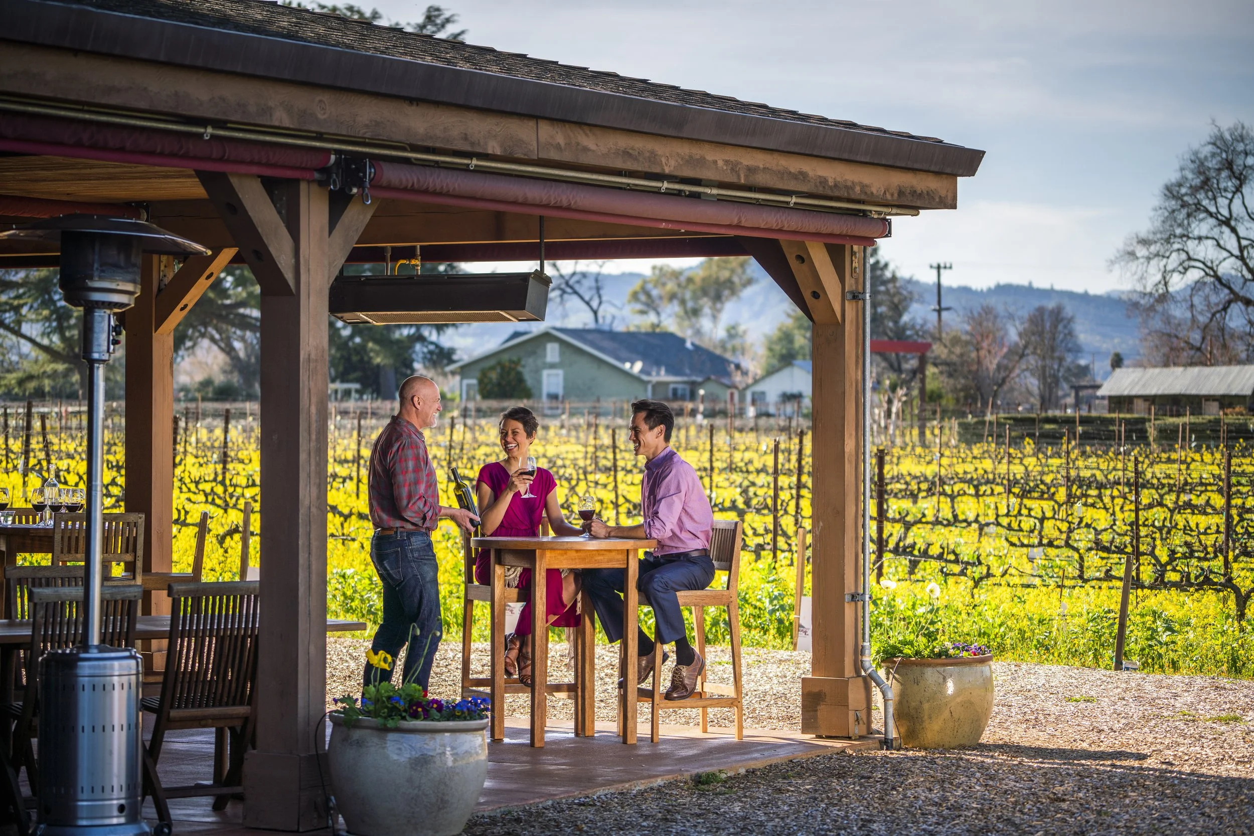 Three people enjoying wine on a patio overlooking a vineyard during sunset.