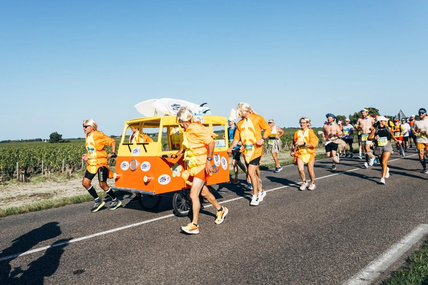 Group of runners dressed in orange and yellow costumes participating in a marathon on a sunny day, with one runner pushing a colorful cart decorated with various stickers and surfboards on top, along a rural road with fields in the background.