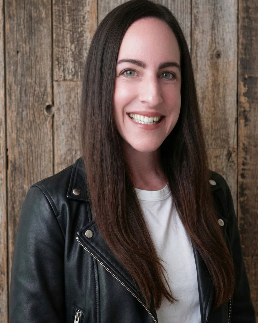 A woman with long dark brown hair, light skin, and blue eyes smiling, wearing a black leather jacket and white T-shirt, standing in front of a wooden wall.