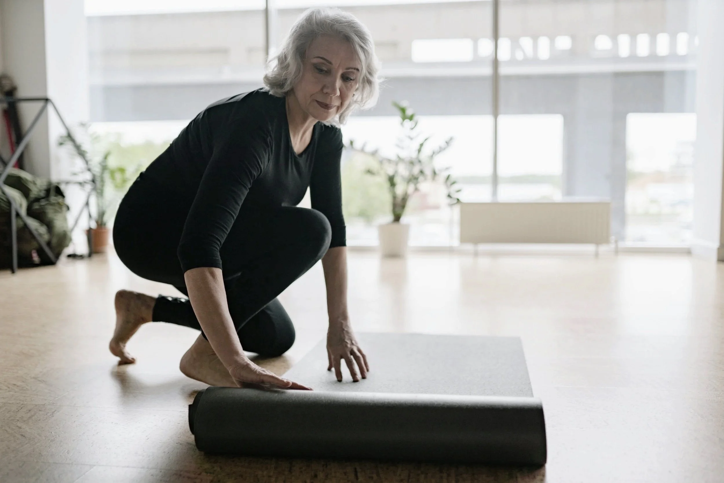 A woman with gray hair in black workout clothes is kneeling on a yoga mat, rolling it out in a bright room with large windows and a plant in the background.