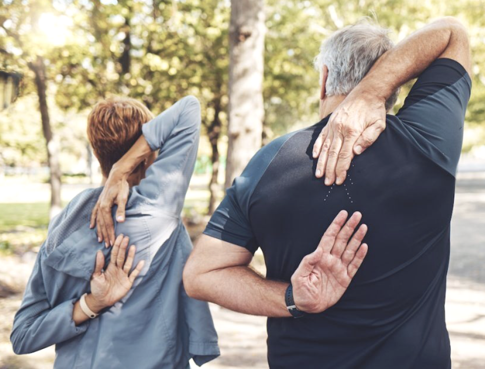 Two elderly people stretching outdoors, facing away from the camera with a background of trees and a park.