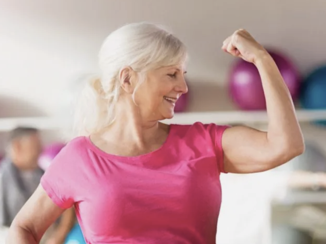 Older woman in a pink shirt flexing her bicep and smiling in a fitness or gym setting.