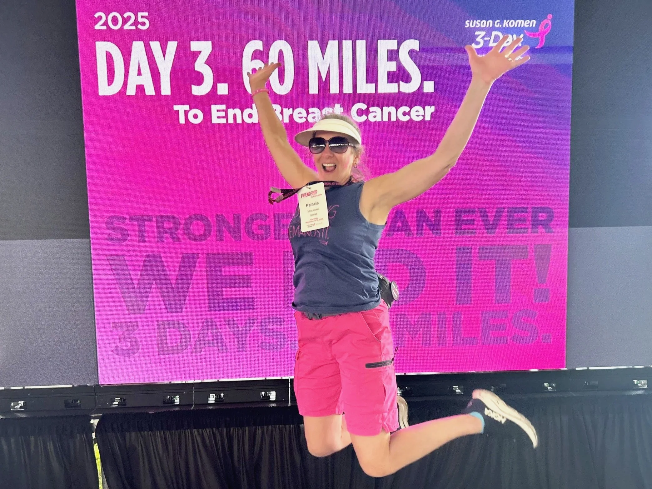 A woman jumping with arms raised in front of a large pink screen displaying the text 'Day 3. 60 miles. To End Breast Cancer' at the Susan G. Komen 3-Day event in 2025. She is wearing sunglasses, a cap, a navy tank top, bright pink shorts, and running shoes.