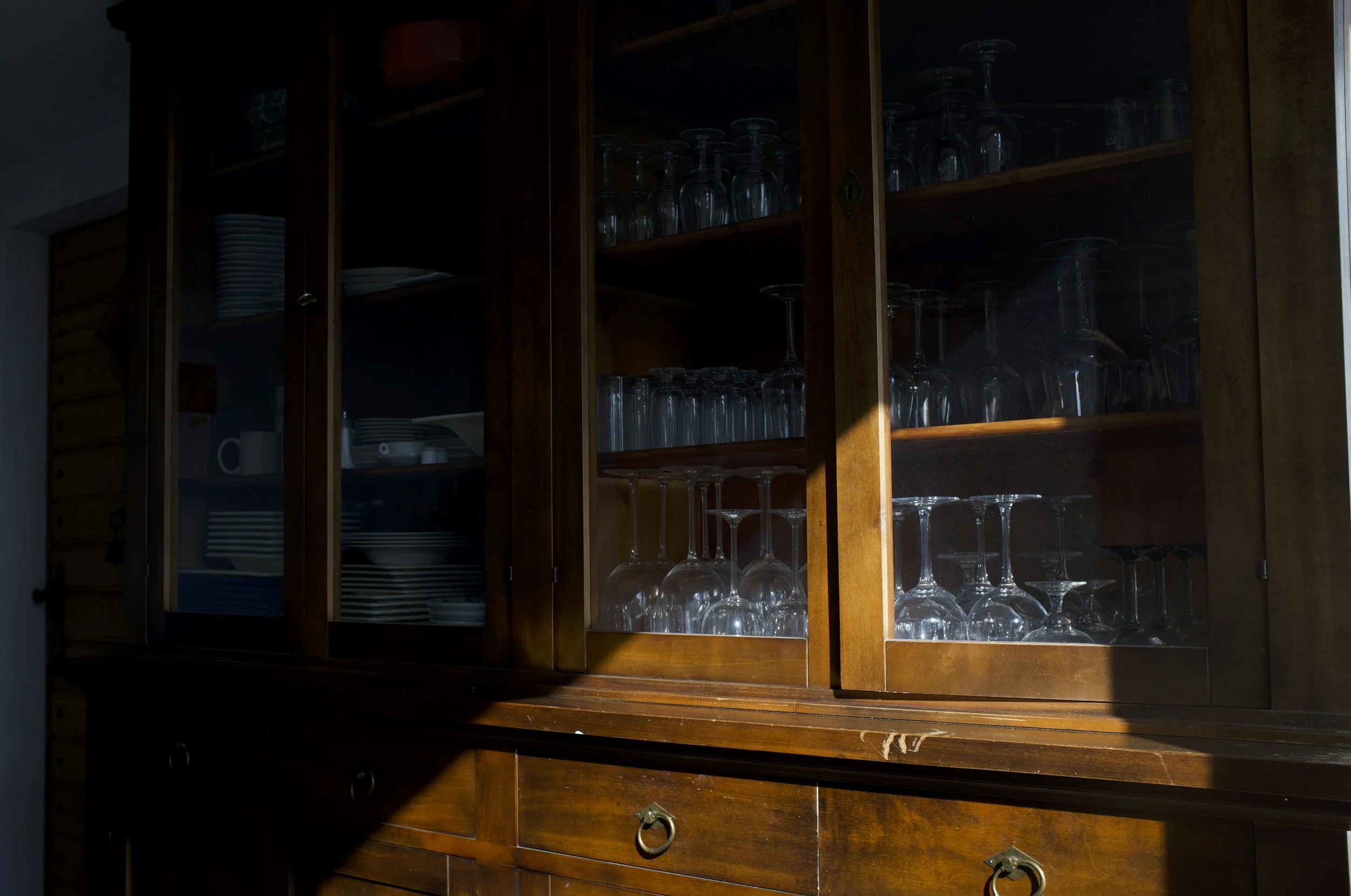 A wooden cabinet with glass doors containing upside-down wine glasses, with sunlight reflecting on the glass and wood surfaces.