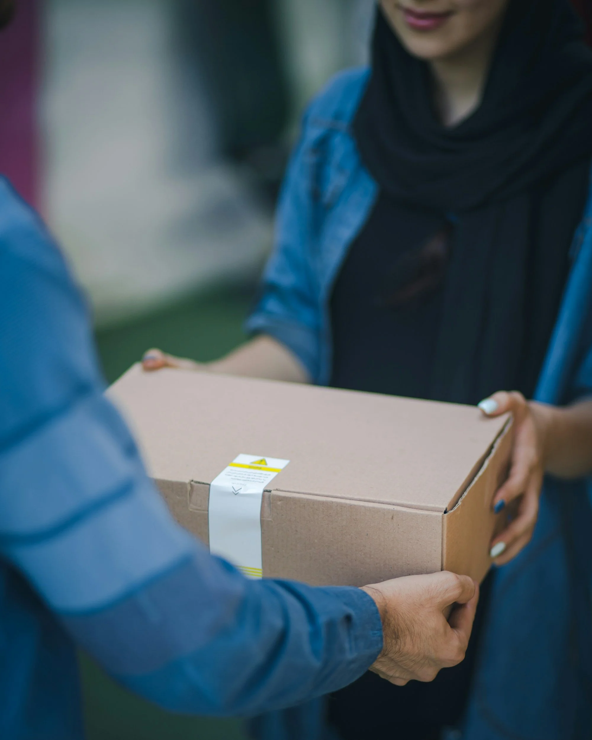 Person in a denim jacket hands a boxed package to another person in a black hoodie.
