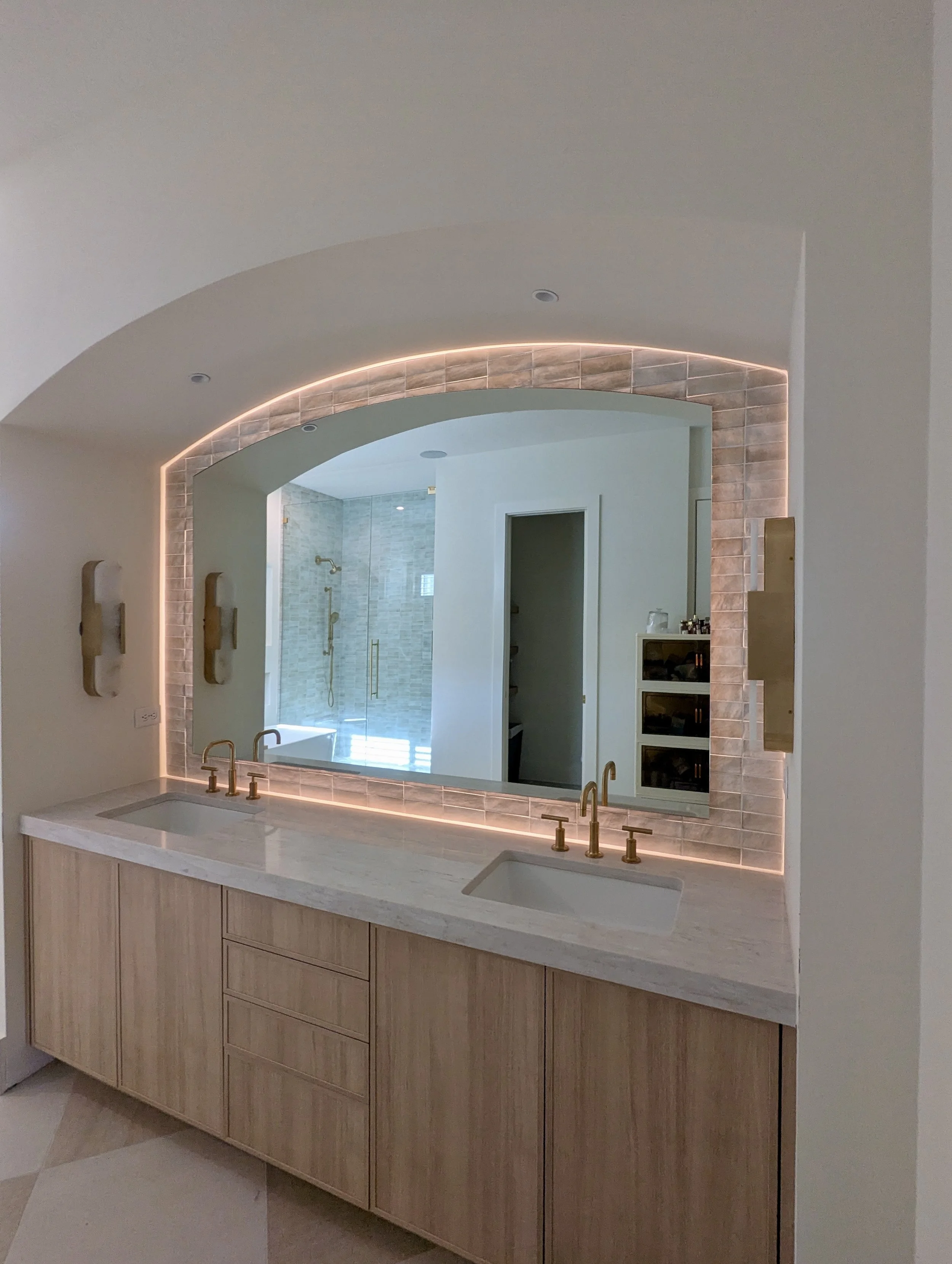 Modern bathroom with double vanity, large backlit mirror, wood cabinets, and a view of a shower with a glass door and gold fixtures.