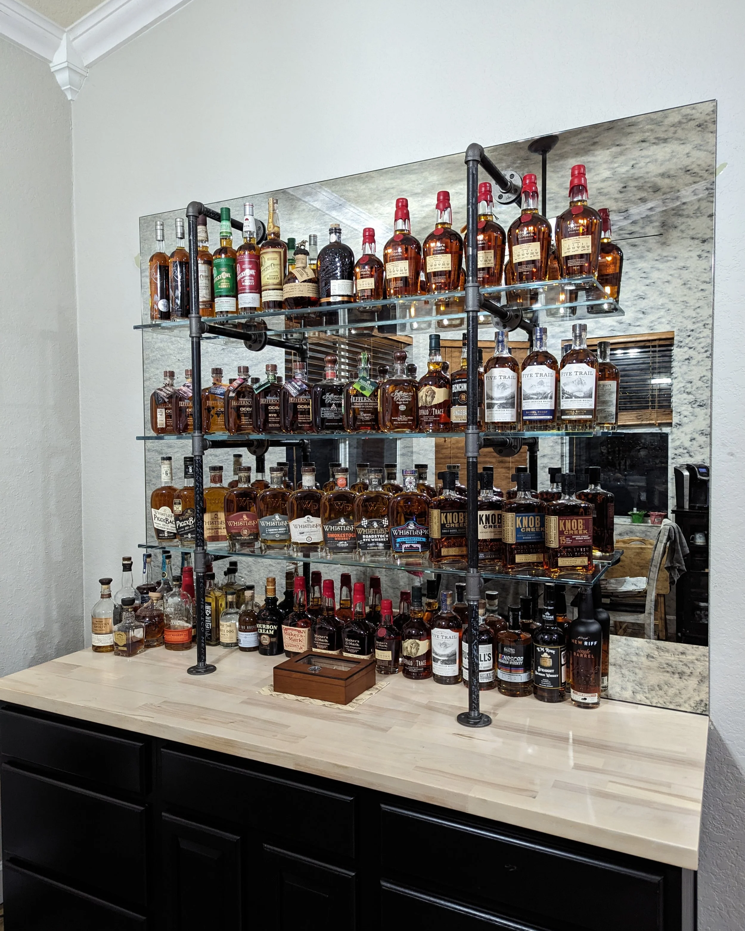 A home bar of various whiskey bottles on glass shelves supported by black piping and a antiqued mirrored wall behind.