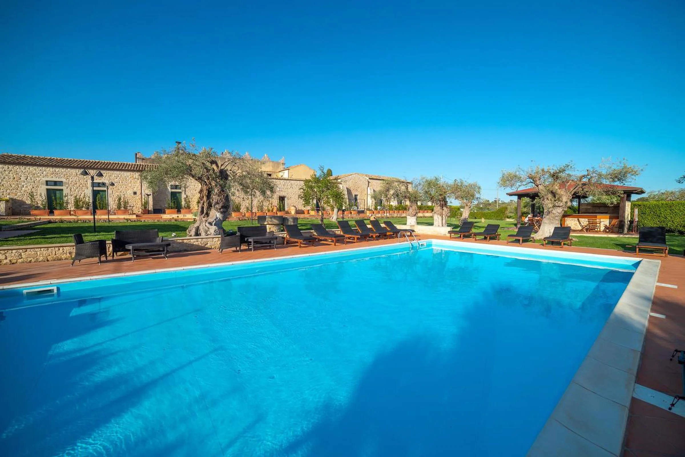 Outdoor swimming pool with lounge chairs, surrounded by trees and stone buildings under a clear blue sky.