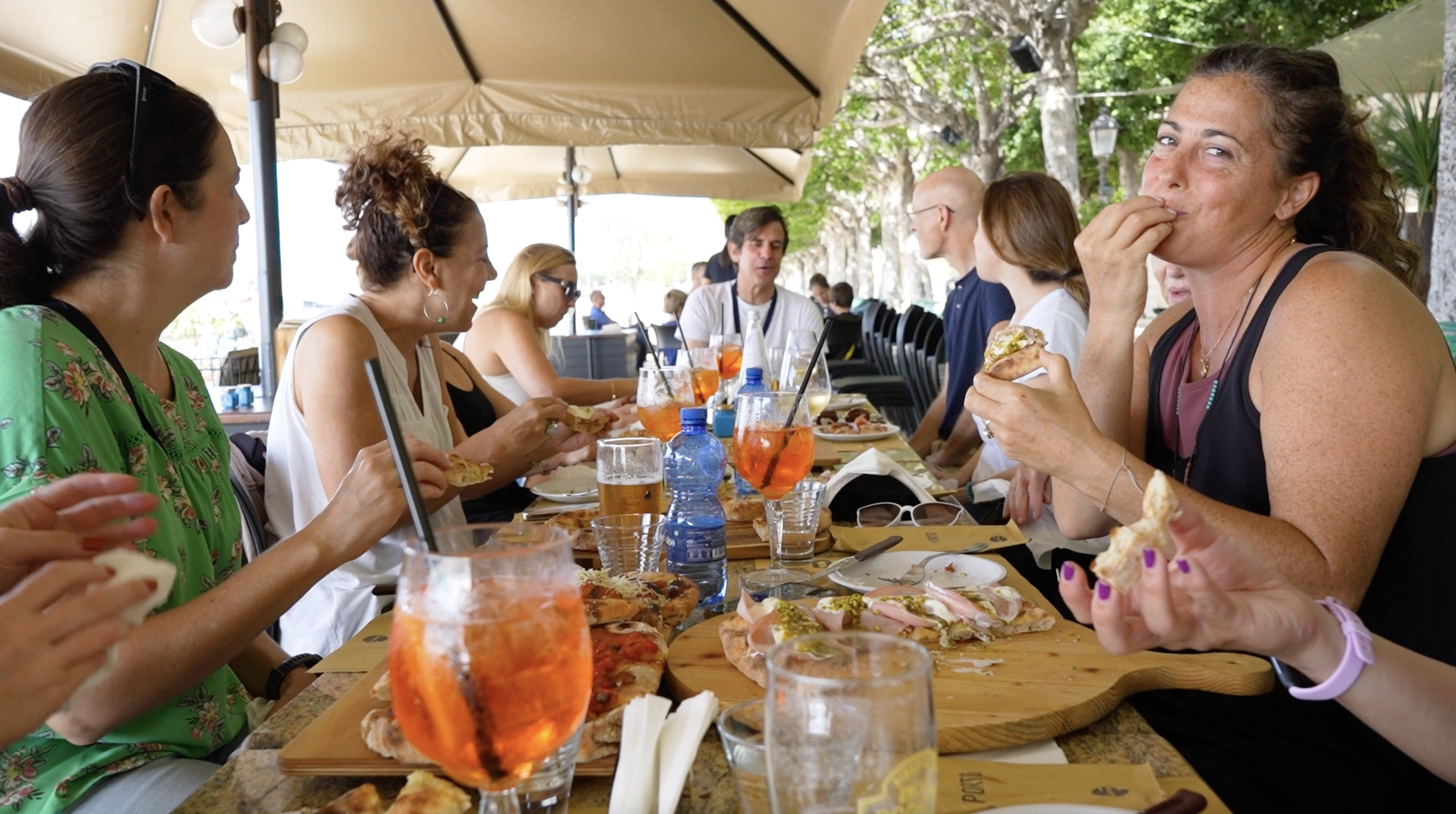 Group of people dining outdoors at a restaurant, enjoying pizza and drinks on a sunny day under umbrellas.