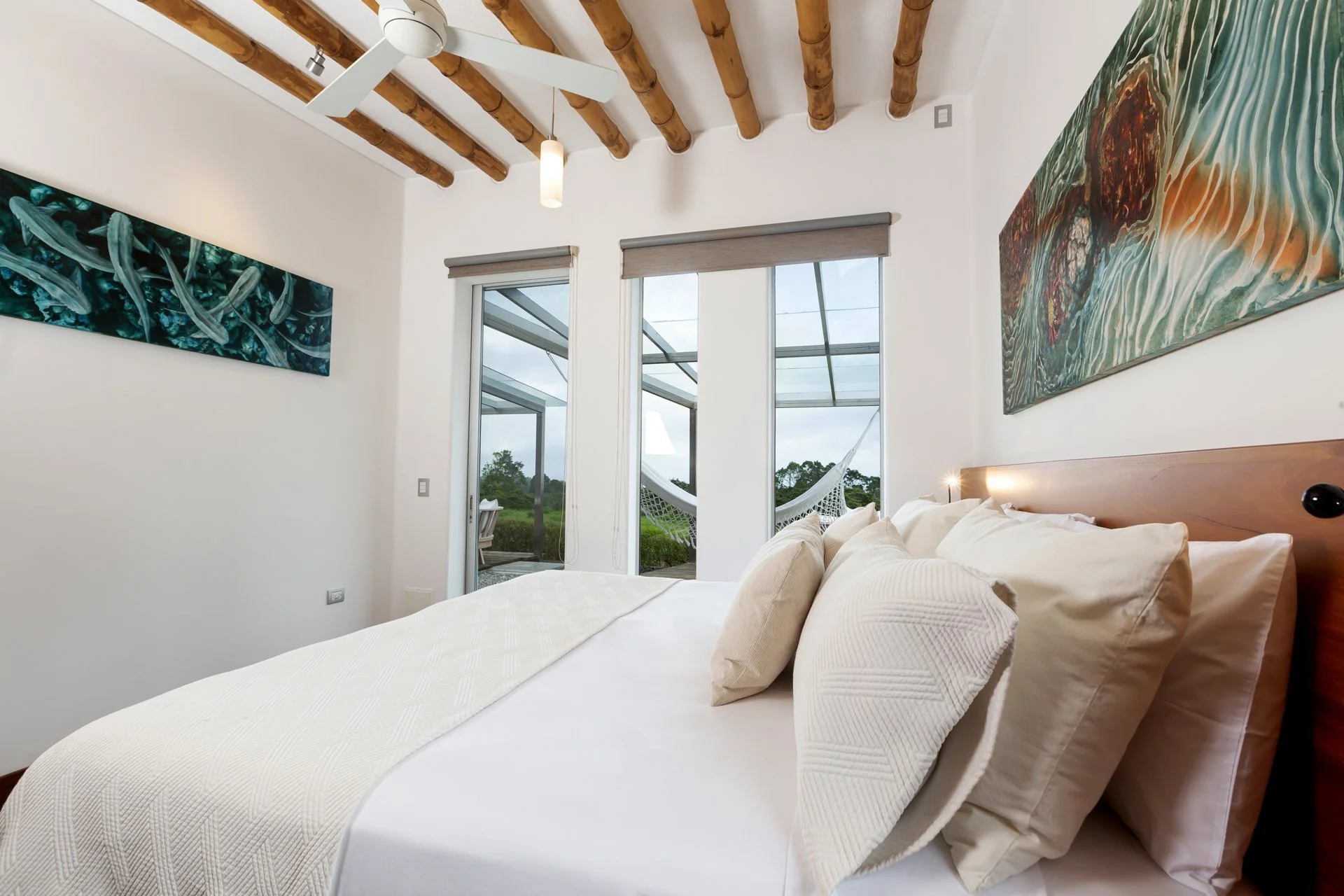 Modern bedroom with white walls, wooden ceiling beams, large windows with views of greenery, a bed with white bedding and pillows, abstract artwork on walls, and a view outside showing a hammock.