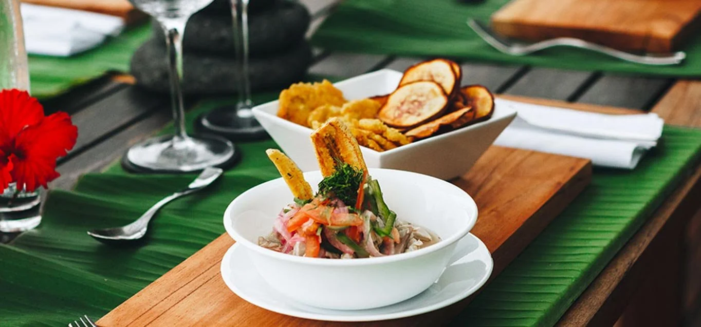 A tropical table setting with a bowl of ceviche, a bowl of potato chips and plantain chips, and two wine glasses on a green banana leaf table runner.