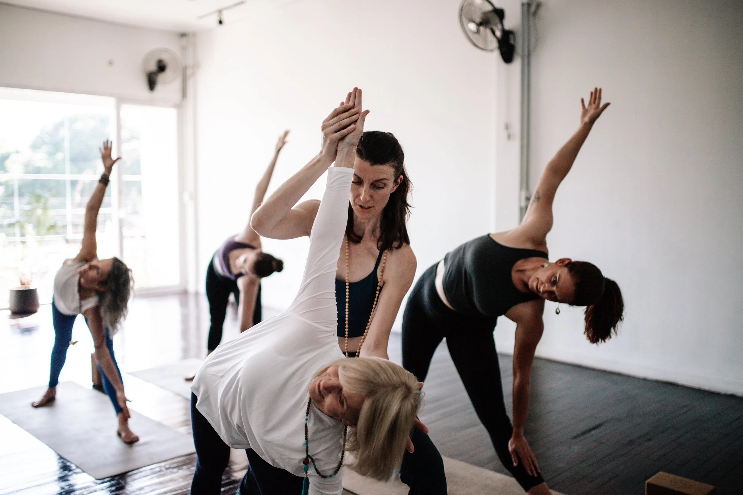 Women practicing yoga in a bright studio with large windows, mats on the floor, and some fans on the wall.