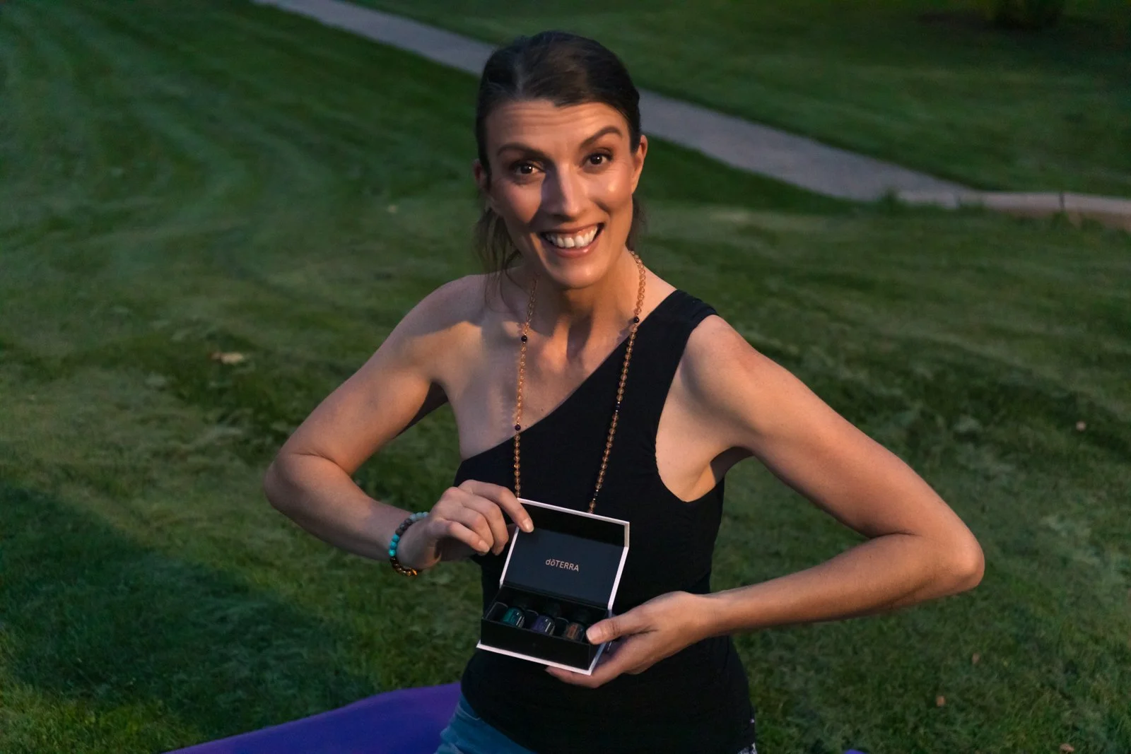A smiling woman outdoors on a grassy area holding a small black box with essential oils, wearing a black sleeveless top and colorful jewelry.