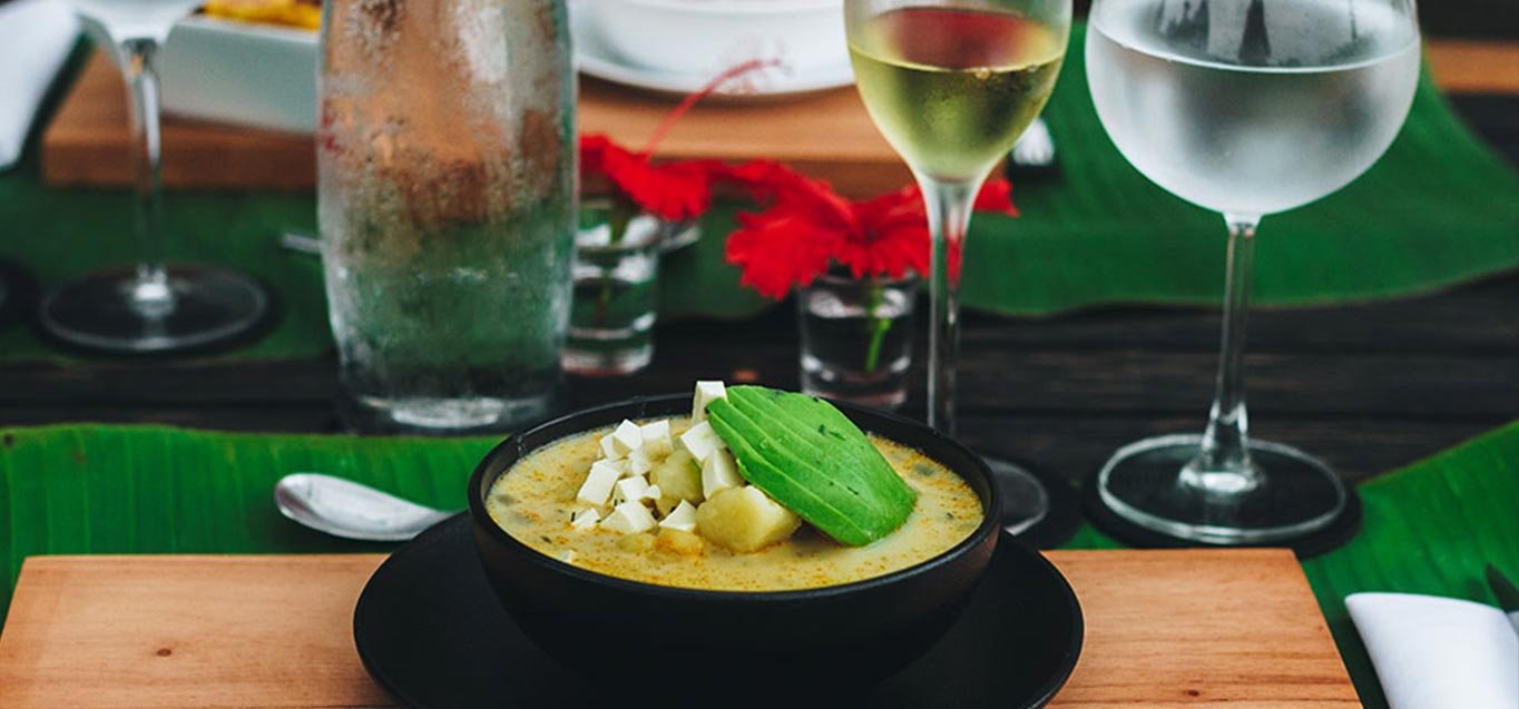 A bowl of soup topped with diced cheese, avocado, and herbs on a wooden table, with glasses of white wine and water in the background.