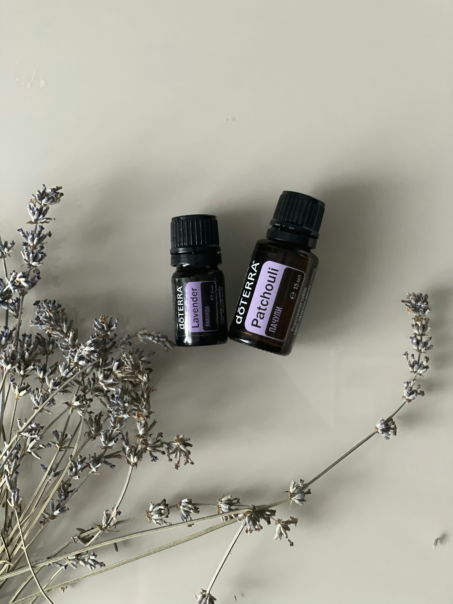Two bottles of doTERRA essential oils, labeled Lavender and Patchouli, placed on a plain white surface with dried lavender flowers around them.