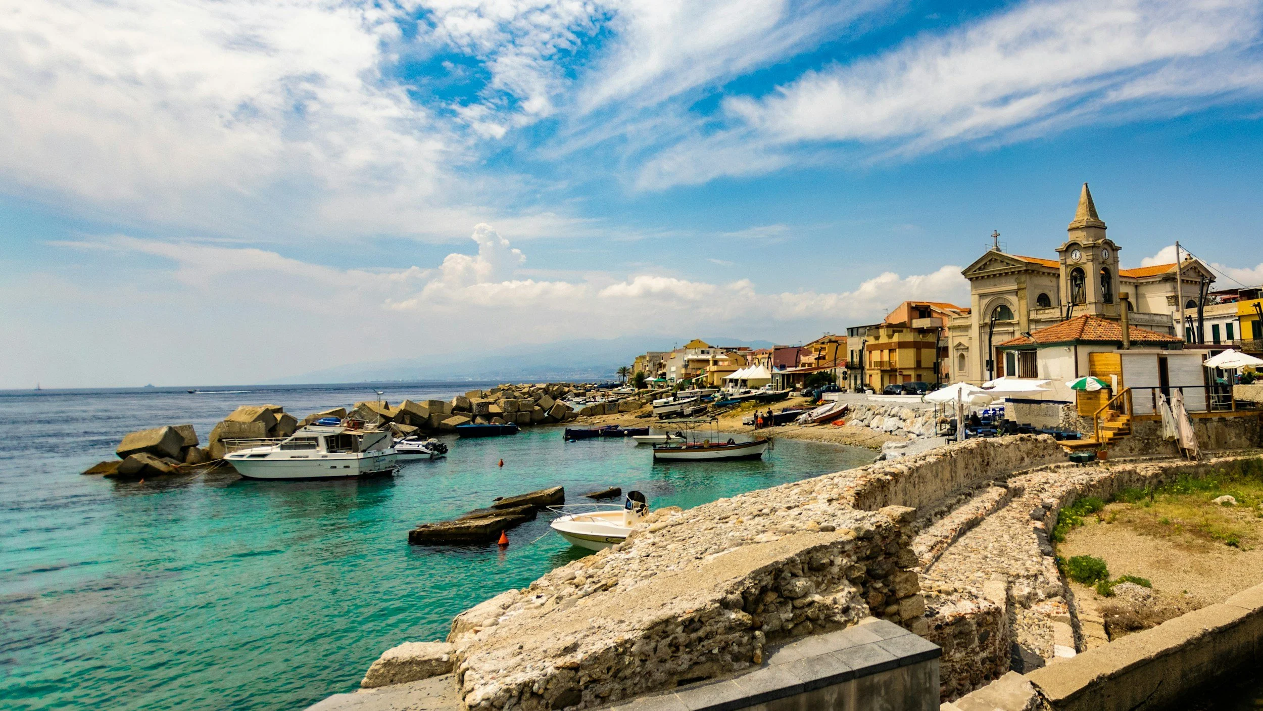 A harbor with boats docked in clear, turquoise water surrounded by rocky breakwaters and a coastal town. The town features colorful buildings, a church with a clock, and a partly cloudy sky.