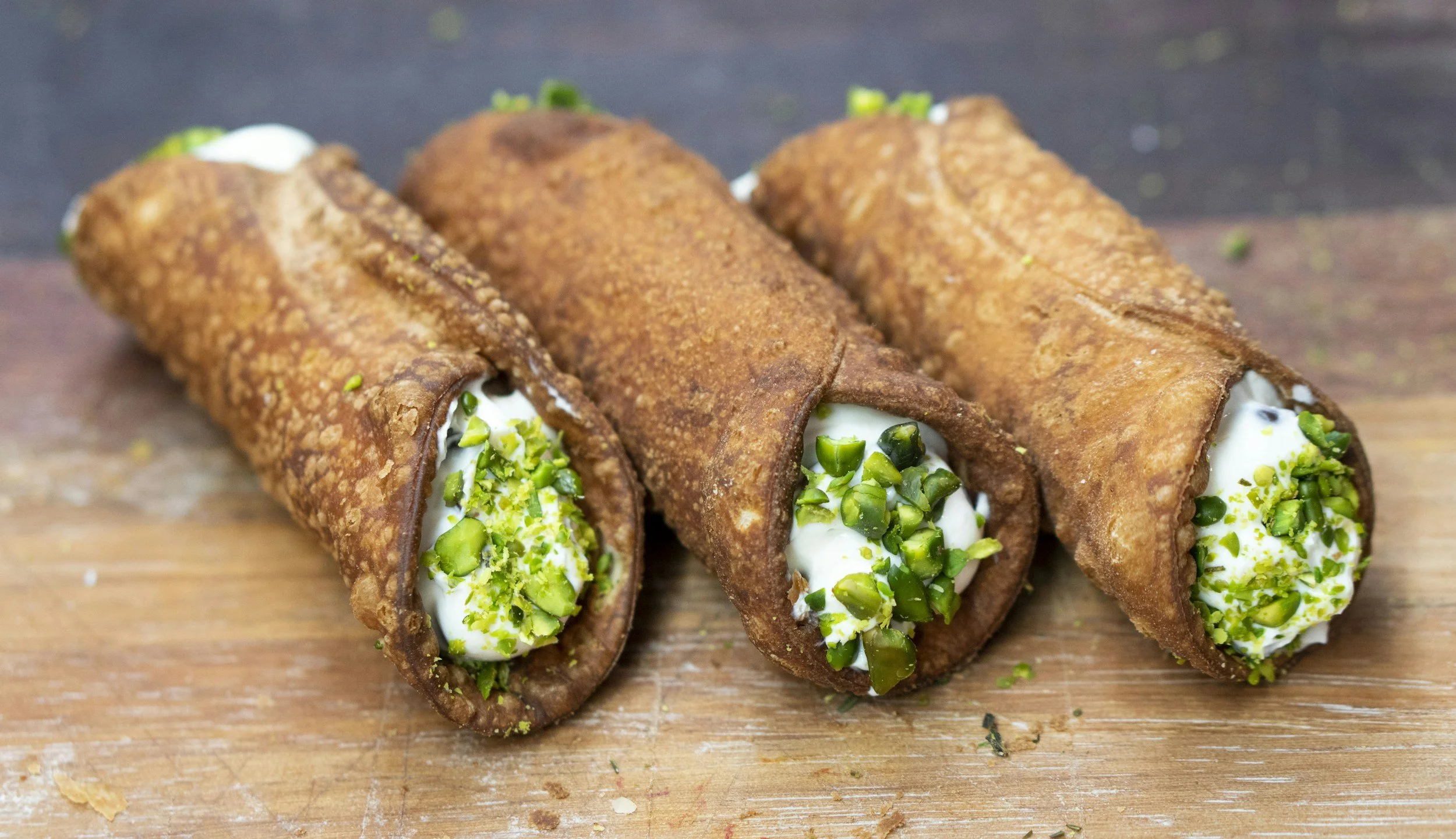 Three cannoli pastries with crispy shell, filled with creamy white filling and topped with chopped pistachios, on a wooden surface.