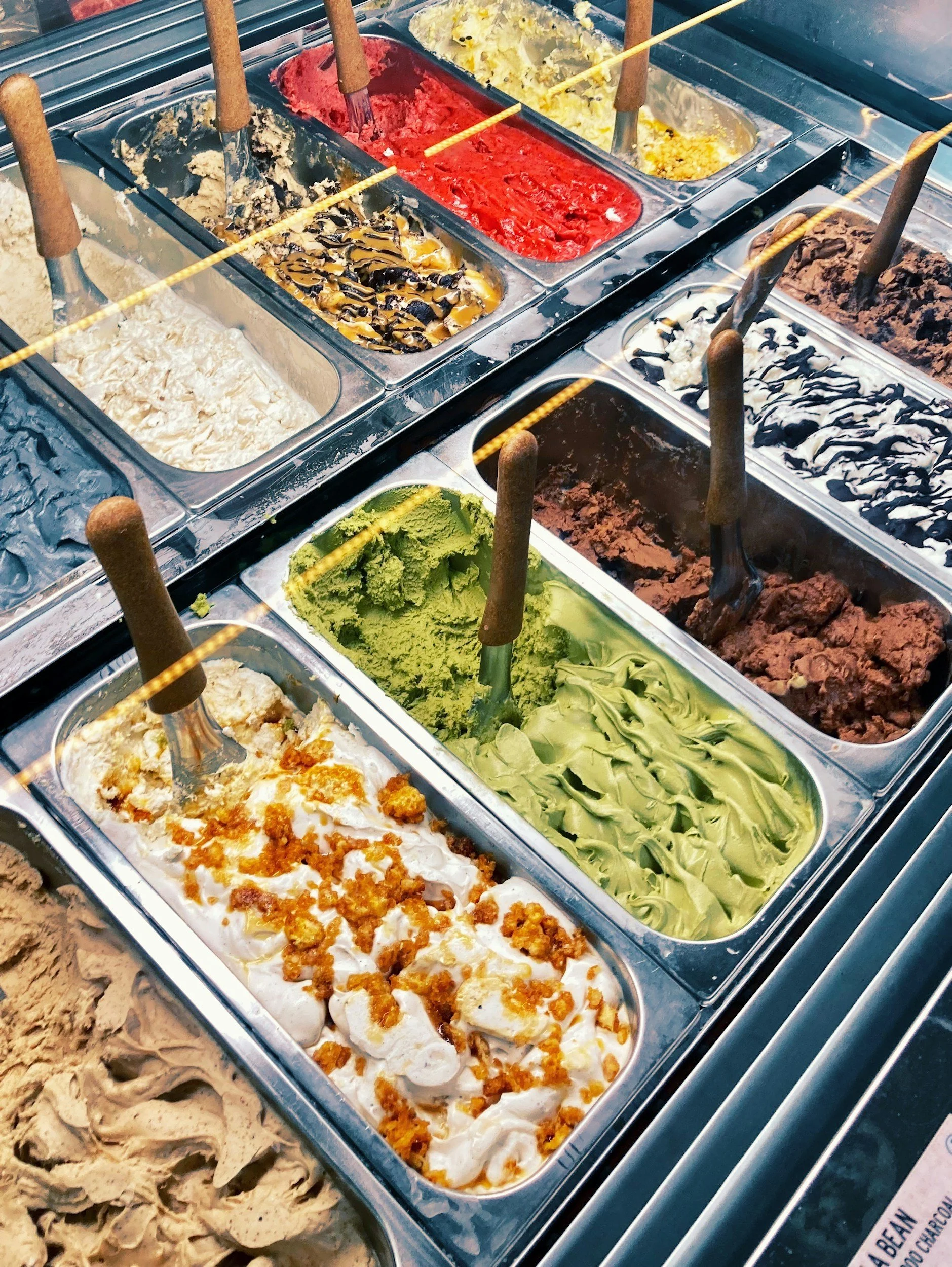 A display case of various flavors of ice cream, including chocolate, green tea, vanilla, strawberry, and others, with scoops and spatulas in each container.