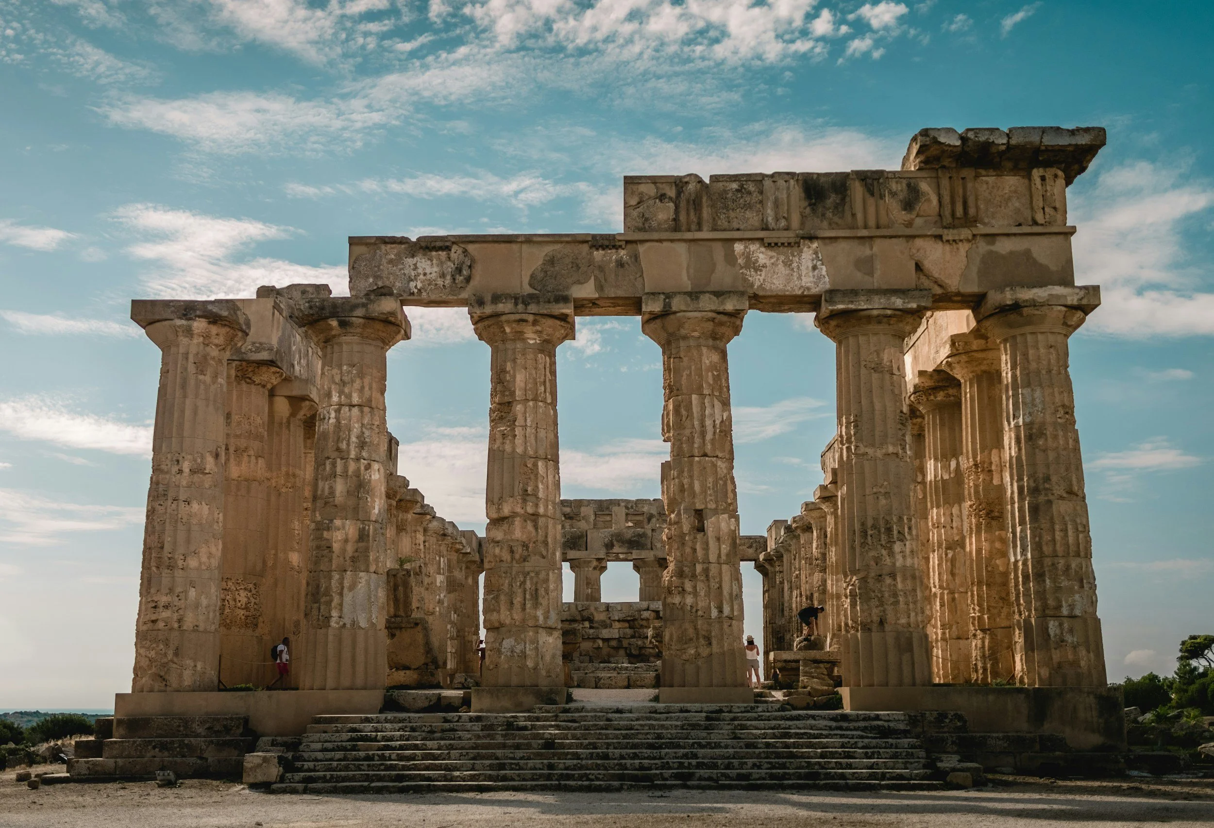 Ancient Greek ruin with tall columns, steps leading up, under a partly cloudy sky.