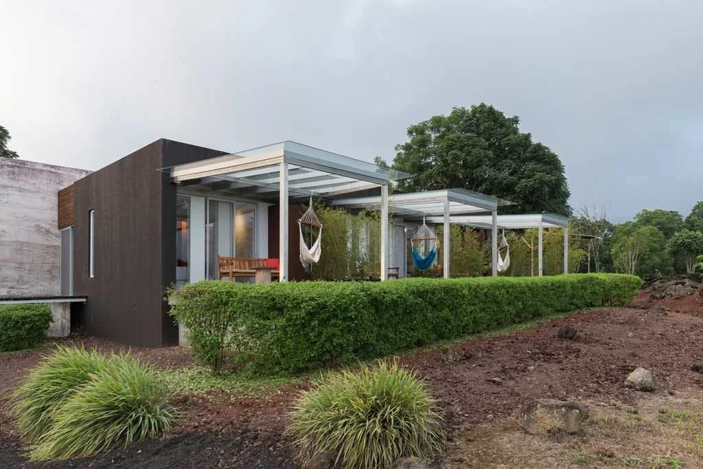 Modern house with covered outdoor lounges and hanging chairs, surrounded by bushes and trees on a cloudy day.