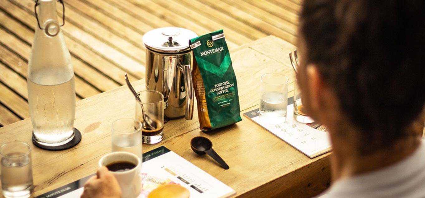 A person sitting at a wooden table with a coffee mug, a water glass, a bottle of water, a bag of game coffee, a spoon, and menu papers.