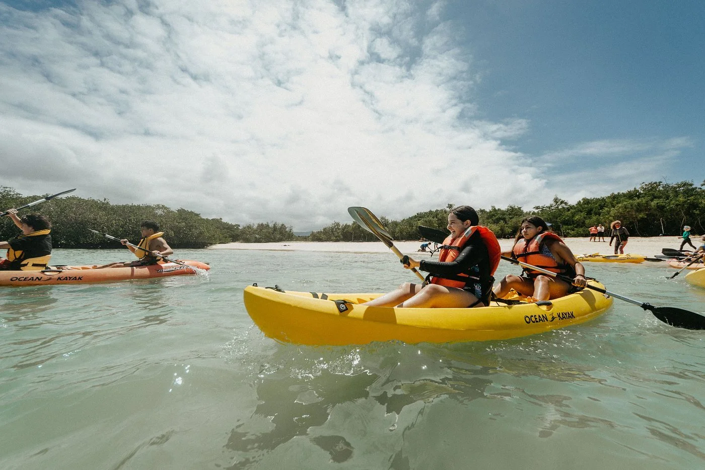 People kayaking in clear water with a beach and cloudy sky in the background.