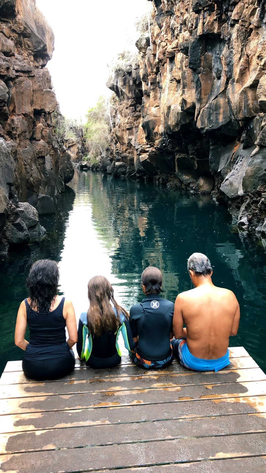 Four people sitting on a wooden dock beside a narrow canyon with rocky walls and calm water.
