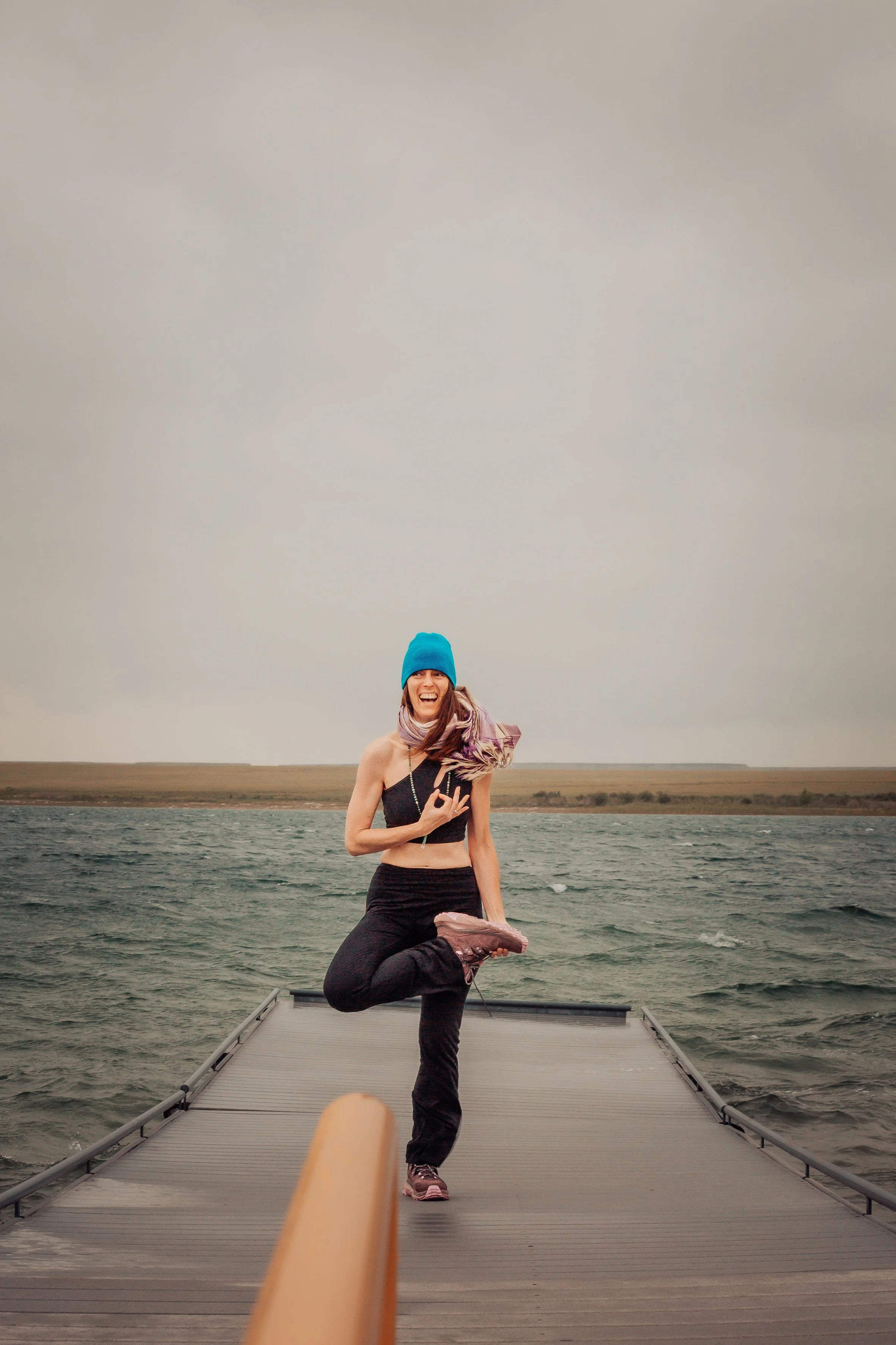 A woman wearing a blue beanie, black crop top, and black pants is standing on one leg on a dock by the water, smiling and making a gesture with hand, with a cloudy sky in the background.