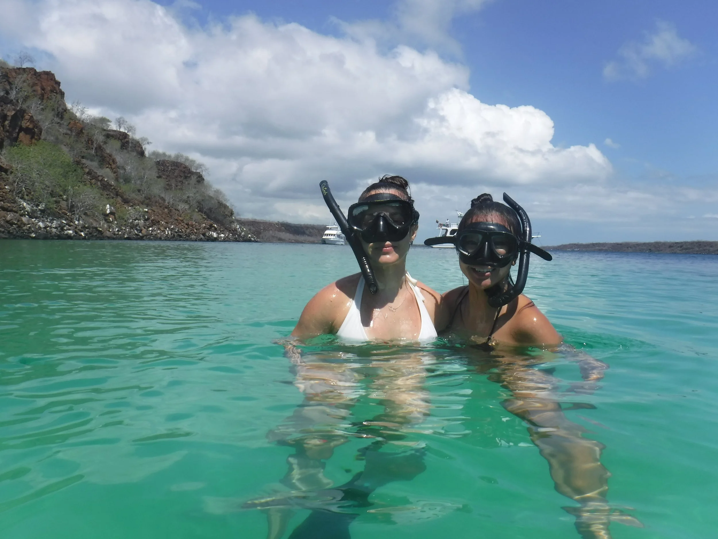 Two women wearing snorkeling masks and swimsuits standing in turquoise water during daytime, with a rocky shoreline, boats, and cloudy sky in the background.