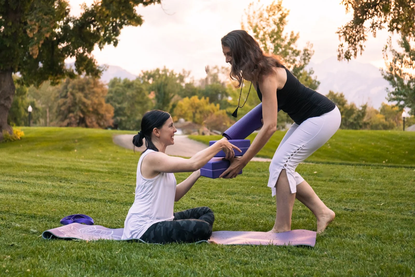 Two women outdoors exchanging yoga mats and blocks, one sitting on a pink yoga mat and the other standing, during sunset in a park with green grass and trees.