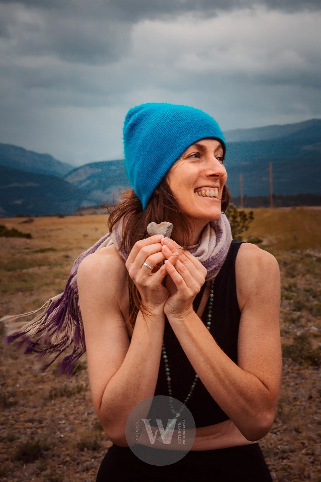 A woman with long brown hair wearing a blue beanie, black sleeveless top, and a purple scarf, holding a small rock and smiling in an open field with mountains and dark clouds in the background.