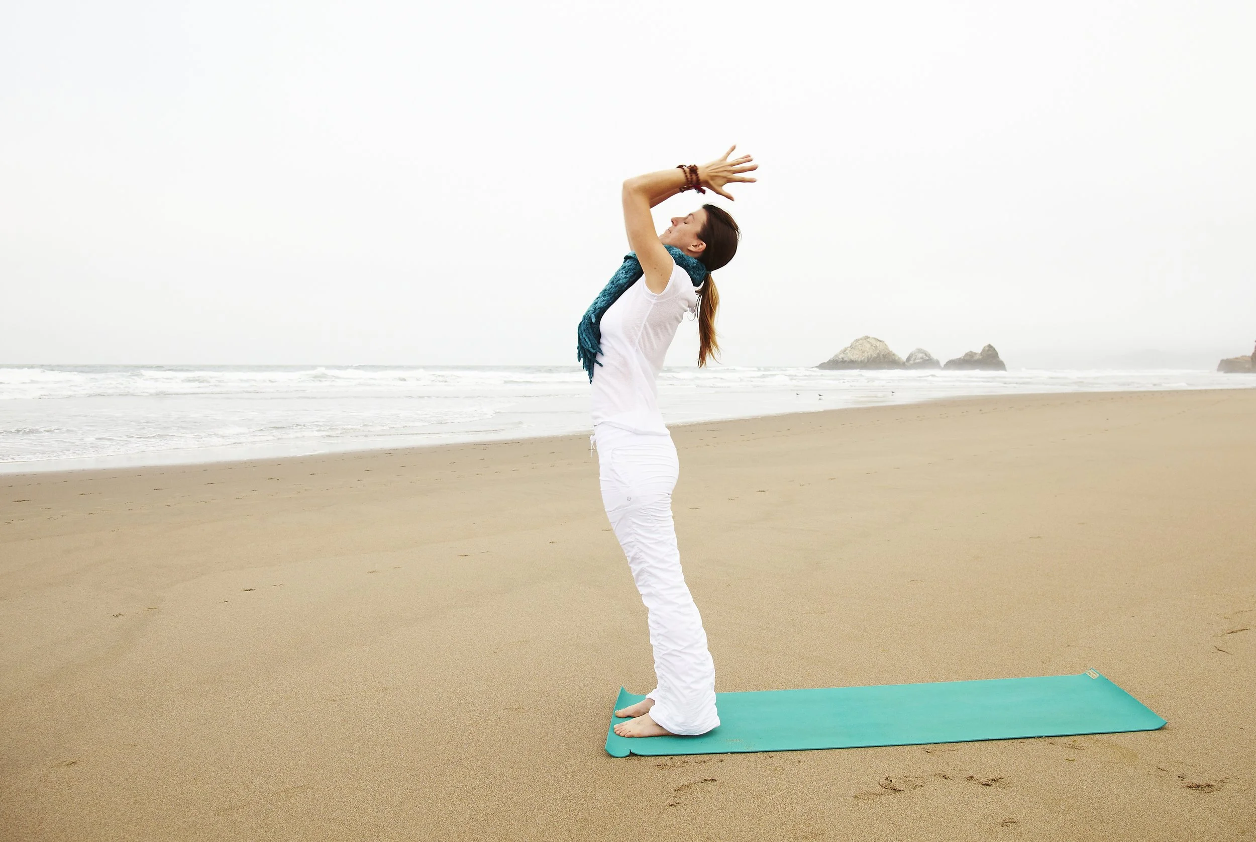 Woman practicing yoga on a beach with ocean waves and rocks in the background.