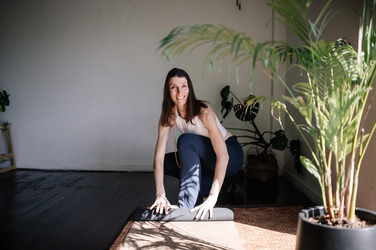 A woman practicing yoga indoors, kneeling on a mat with a yoga roll, surrounded by houseplants, smiling at the camera.