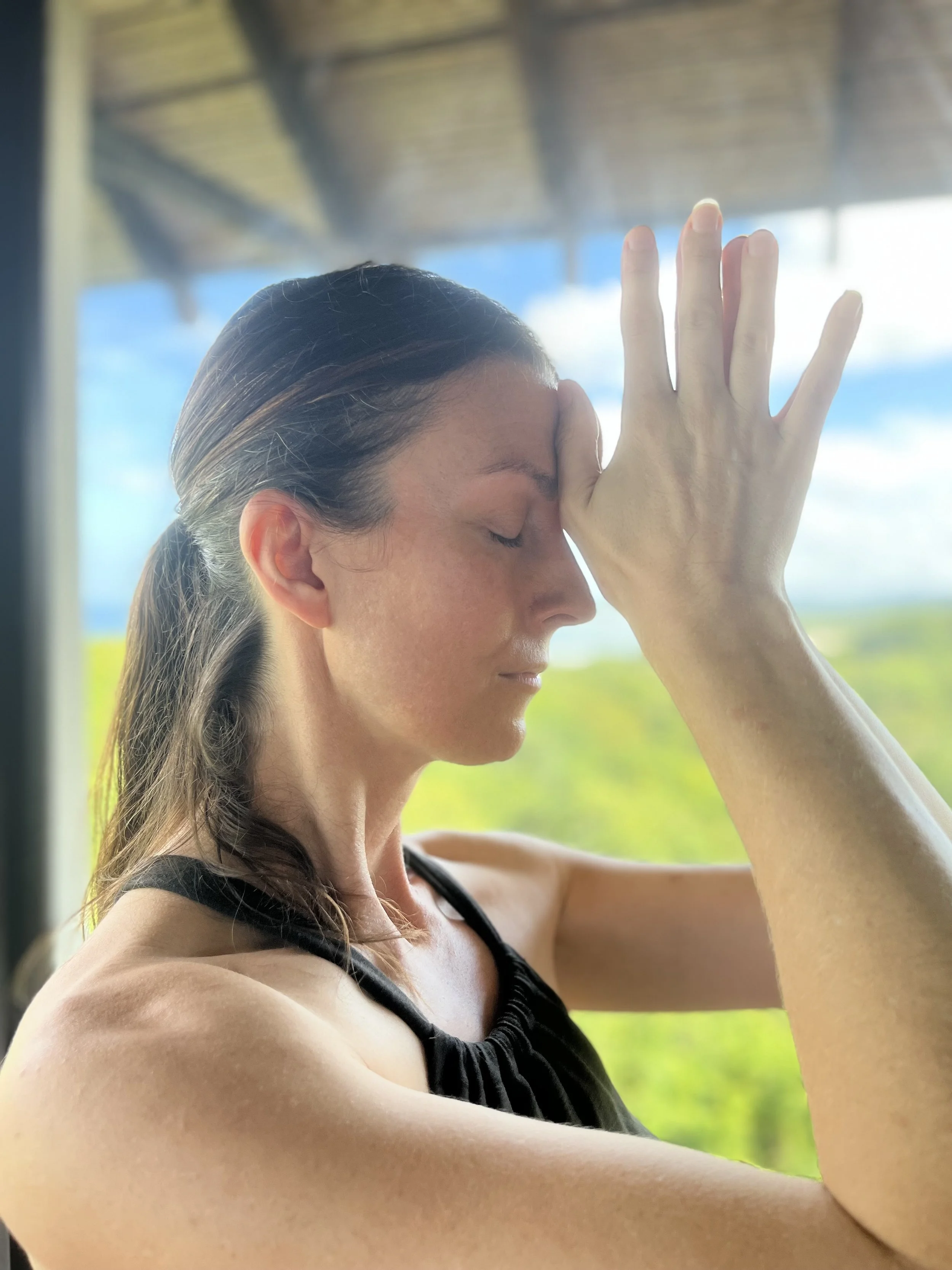 Woman practicing yoga or meditation indoors with a serene expression, eyes closed, hand resting on her forehead, background showing a bright outdoor scene with trees.