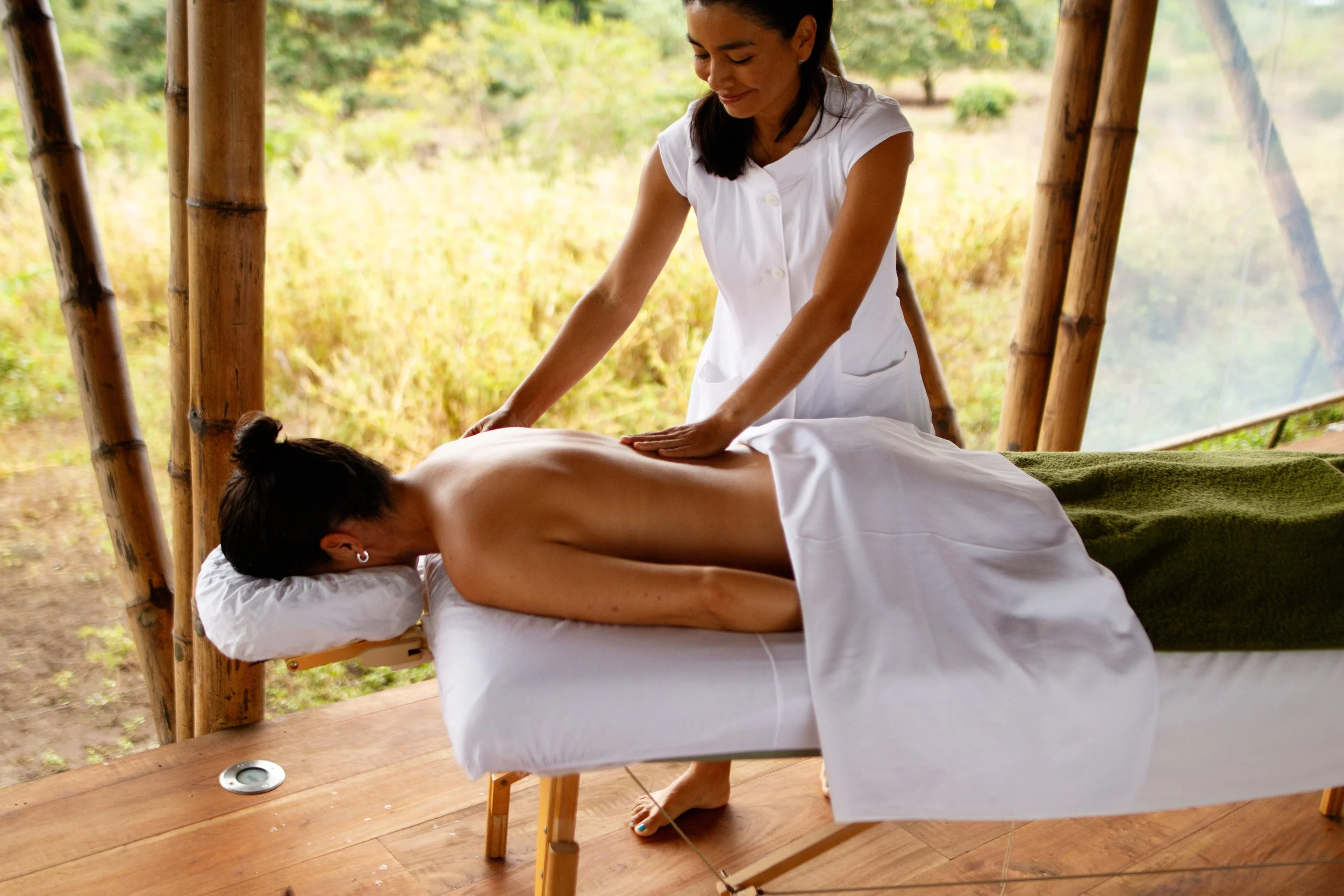 A woman receives a back massage from a massage therapist in an outdoor setting with trees and bamboo in the background.