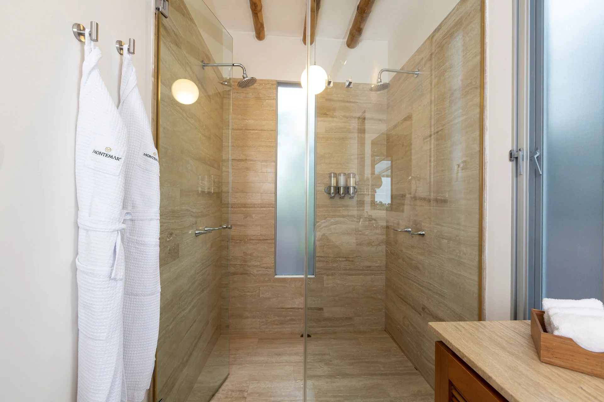 A modern bathroom with a glass-enclosed shower featuring beige marble tiles, two showerheads, and a frosted window. Two white towels hang on hooks, and a wooden countertop with rolled white towels is visible.