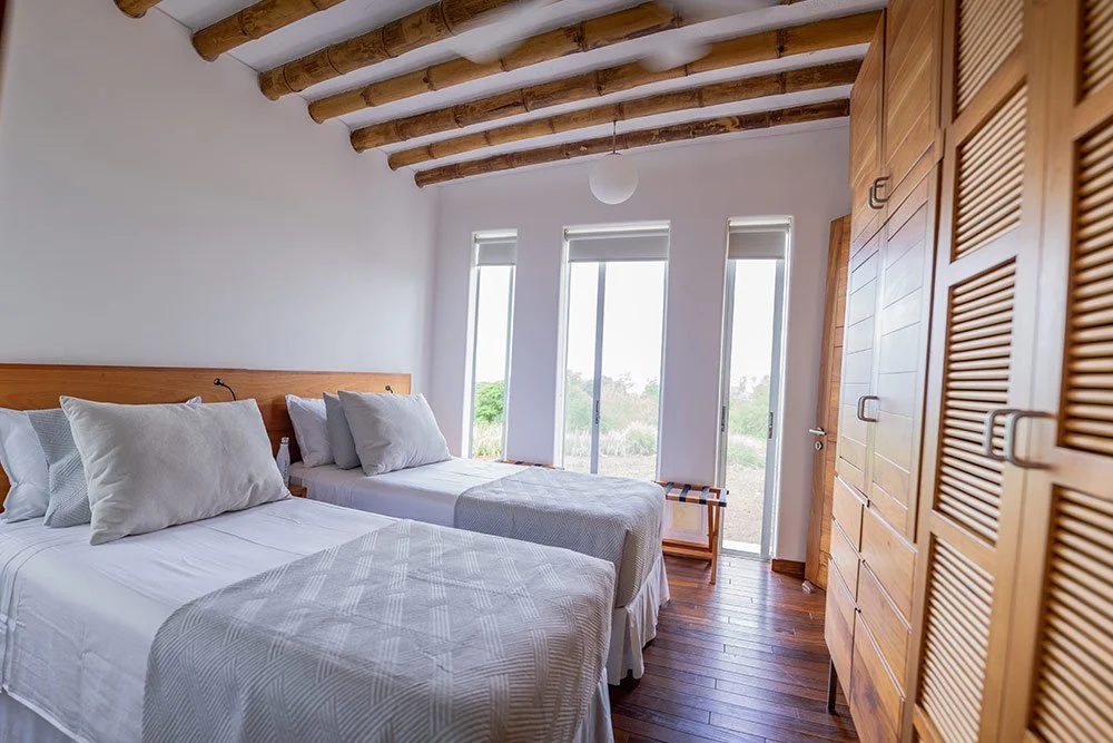 A bedroom with two twin beds with white bedding and pillows, wooden ceiling beams, large windows with a view of greenery, and wooden furniture including a wardrobe and side table.
