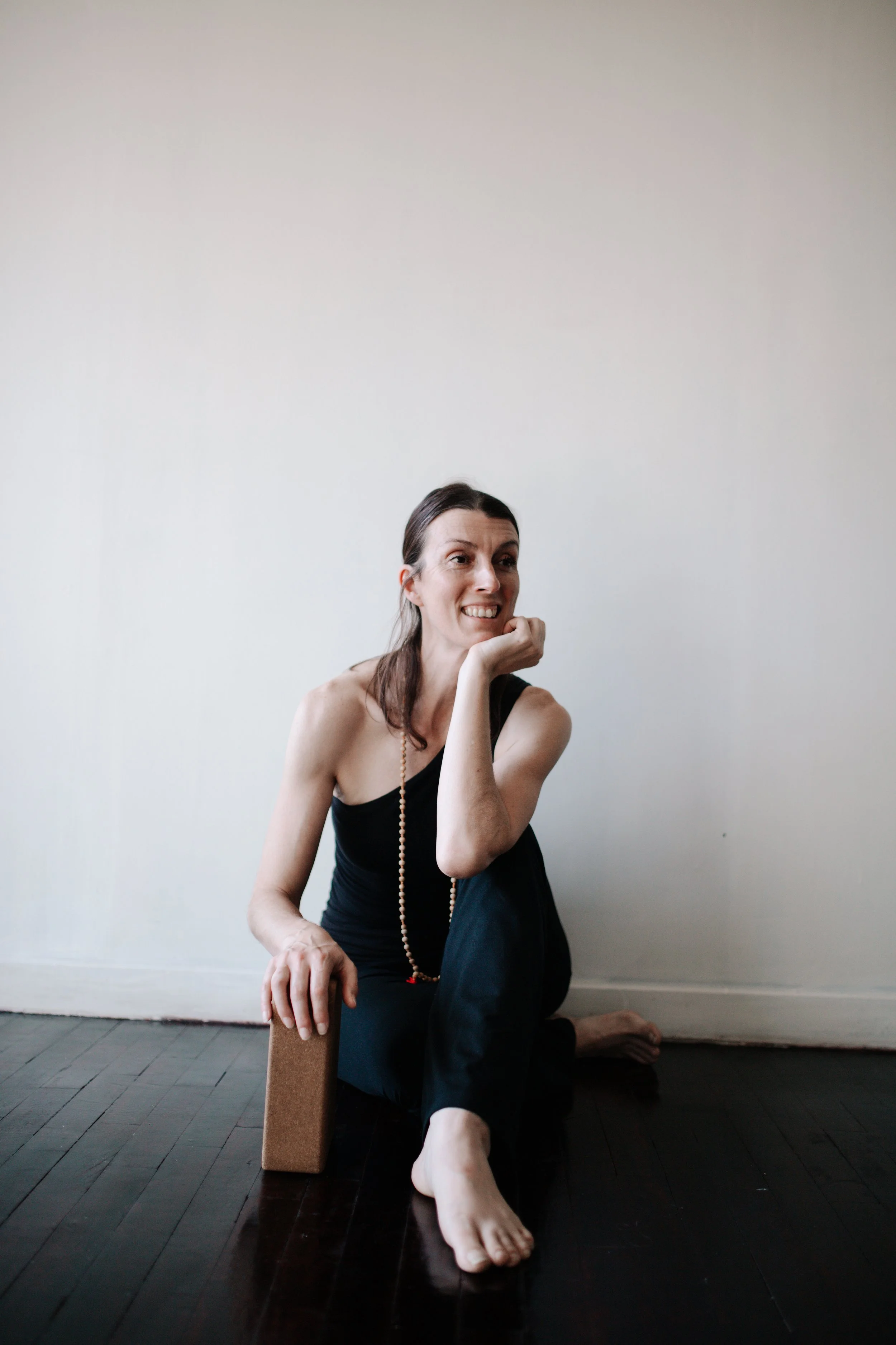 A woman sits on a dark wooden floor, smiling and resting her chin on her right hand, with her left hand on a cork yoga block. She is wearing a black sleeveless top, black pants, and a beaded necklace, against a plain white wall.