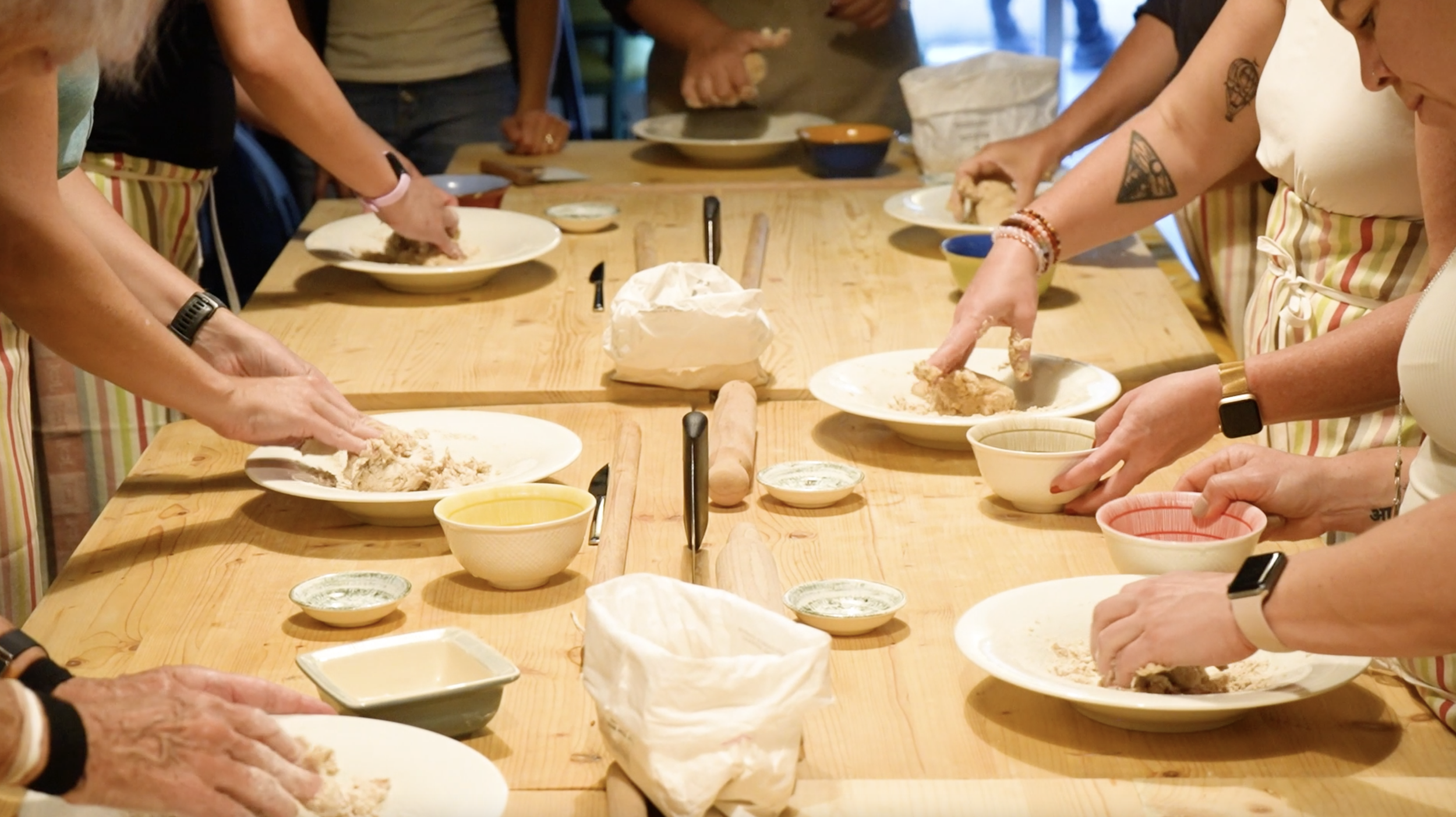 People gathered around a wooden table, preparing dough with their hands while wearing aprons.