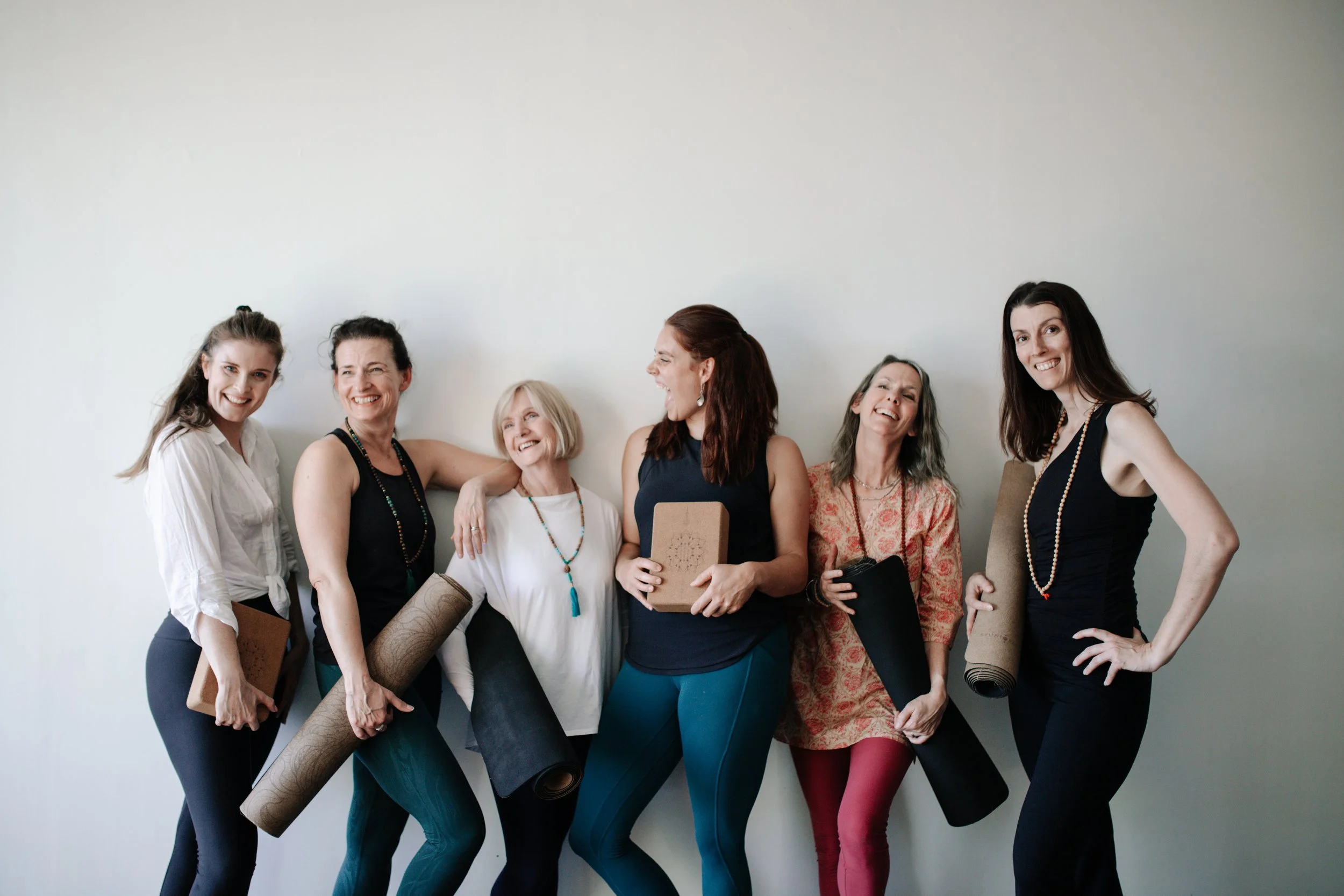 Six women standing against a white wall, smiling, holding yoga mats and books, dressed in casual athletic attire.