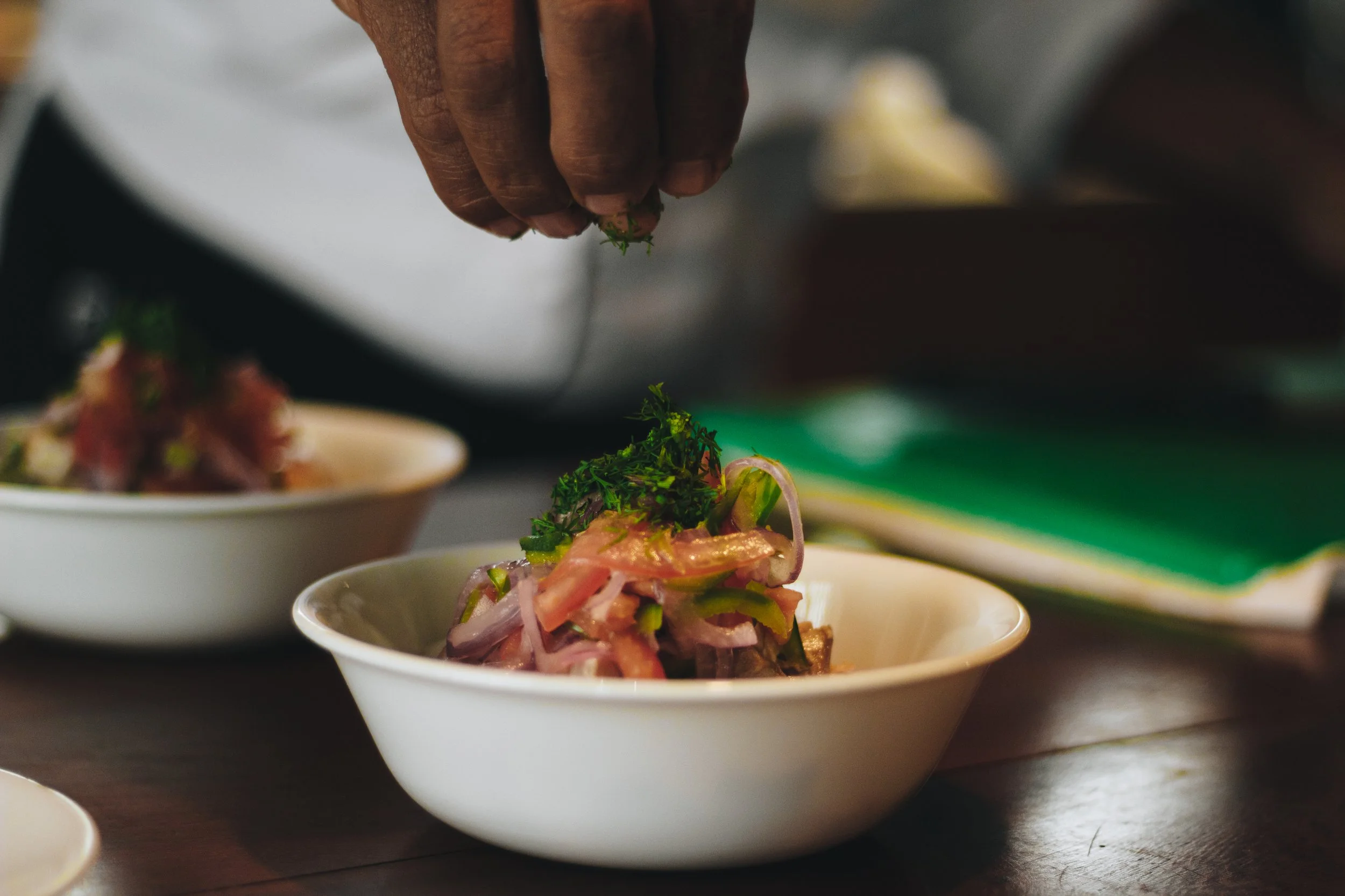A hand sprinkling chopped herbs onto a bowl of colorful salad with thinly sliced vegetables on a wooden table.